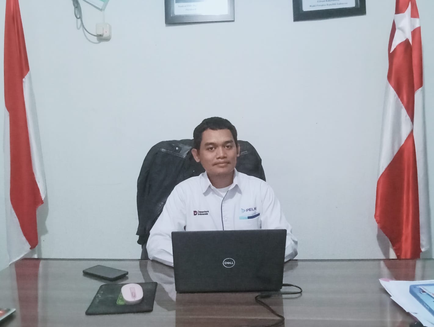 Pejabat Dinas Perhubungan Kepulauan Aru duduk di meja kerja dengan laptop dan bendera merah putih di belakangnya, menggambarkan suasana resmi saat membahas kesiapan KM Labobar dan surat Pemkab kepada Dirjen Hubla.