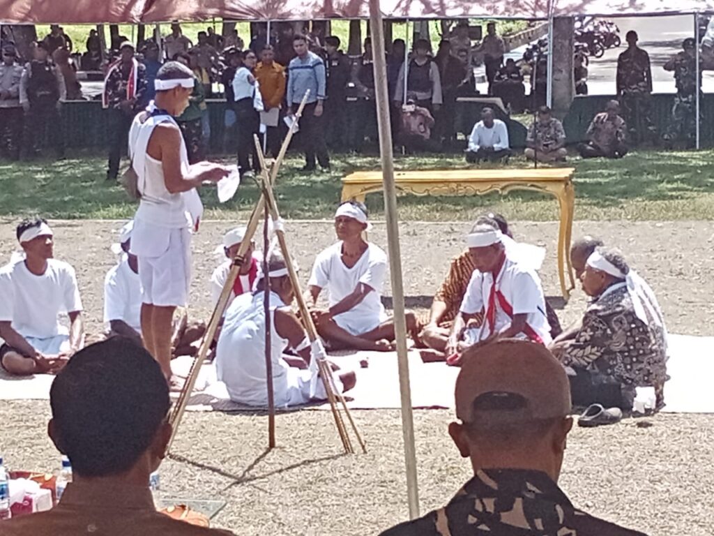 okoh adat dari kedua desa duduk melingkar di bawah tenda, mengenakan pakaian putih dan ikat kepala, melaksanakan ritual adat perdamaian.