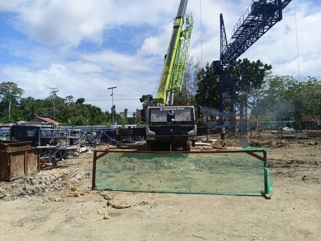 Aktivitas proyek RSUD Dobo menampilkan crane hijau dan struktur baja di area konstruksi yang dikelilingi pepohonan, menggambarkan proses bongkar muat material oleh pekerja lapangan.