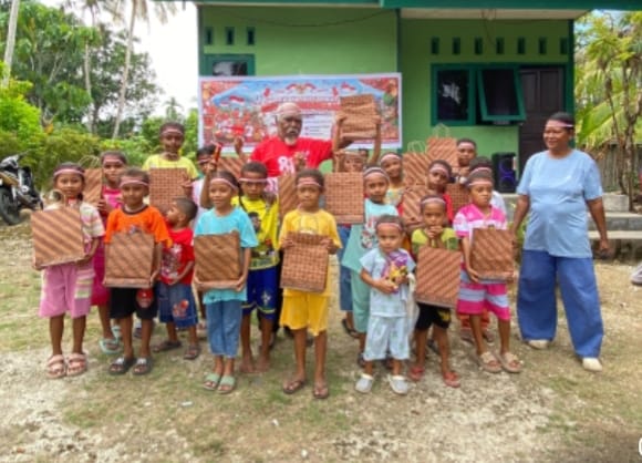 Anak-anak Papua berbaris sambil memegang hadiah anyaman tradisional di depan bangunan hijau dengan spanduk HUT RI ke-81.