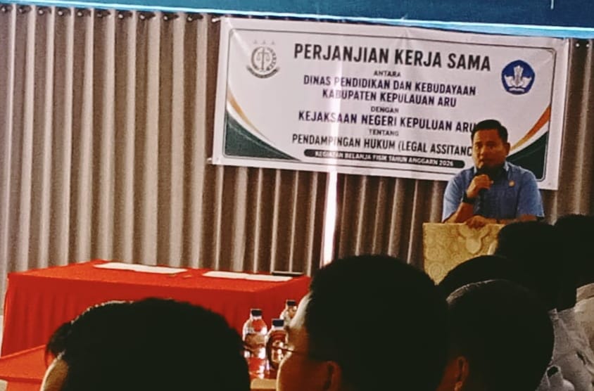Kajari Kepulauan Aru berbicara di podium di depan peserta sosialisasi perjanjian kerja sama pendampingan hukum dengan latar spanduk resmi kegiatan.