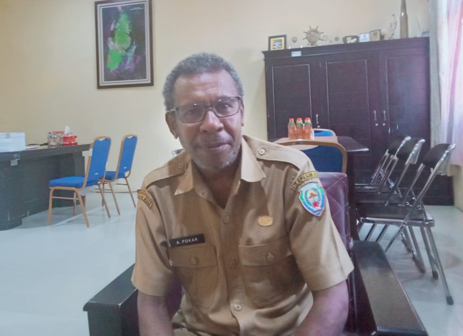 Kepala Dinas Pendidikan Kepulauan Aru mengenakan seragam dinas cokelat duduk di ruang kerja dengan latar meja dan kursi kantor.
