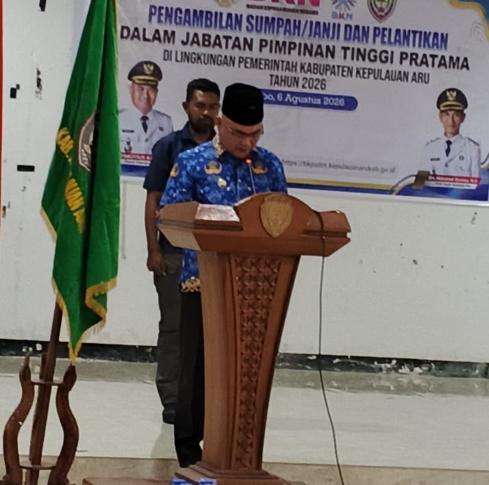 Pejabat mengenakan batik biru berdiri di podium membacakan sumpah jabatan dengan latar spanduk pelantikan di aula Pemkab Kepulauan Aru.