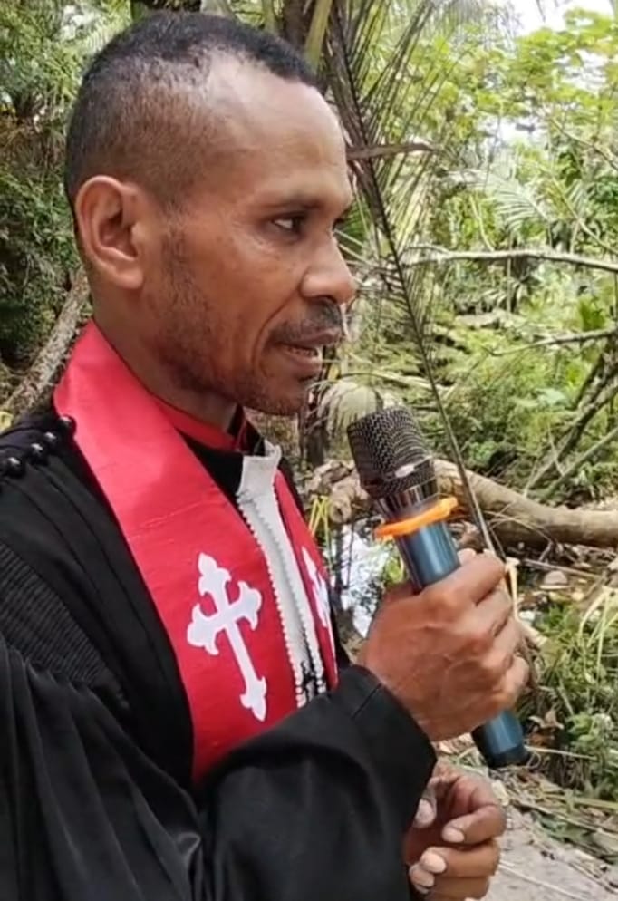 Seorang pendeta pria GKI dari suku asli Papua memberikan khotbah dengan mikrofon pada ibadah HUT ke-22 GKI Imanuel Nyiben, mengenakan jubah hitam dan stola merah dengan salib putih.