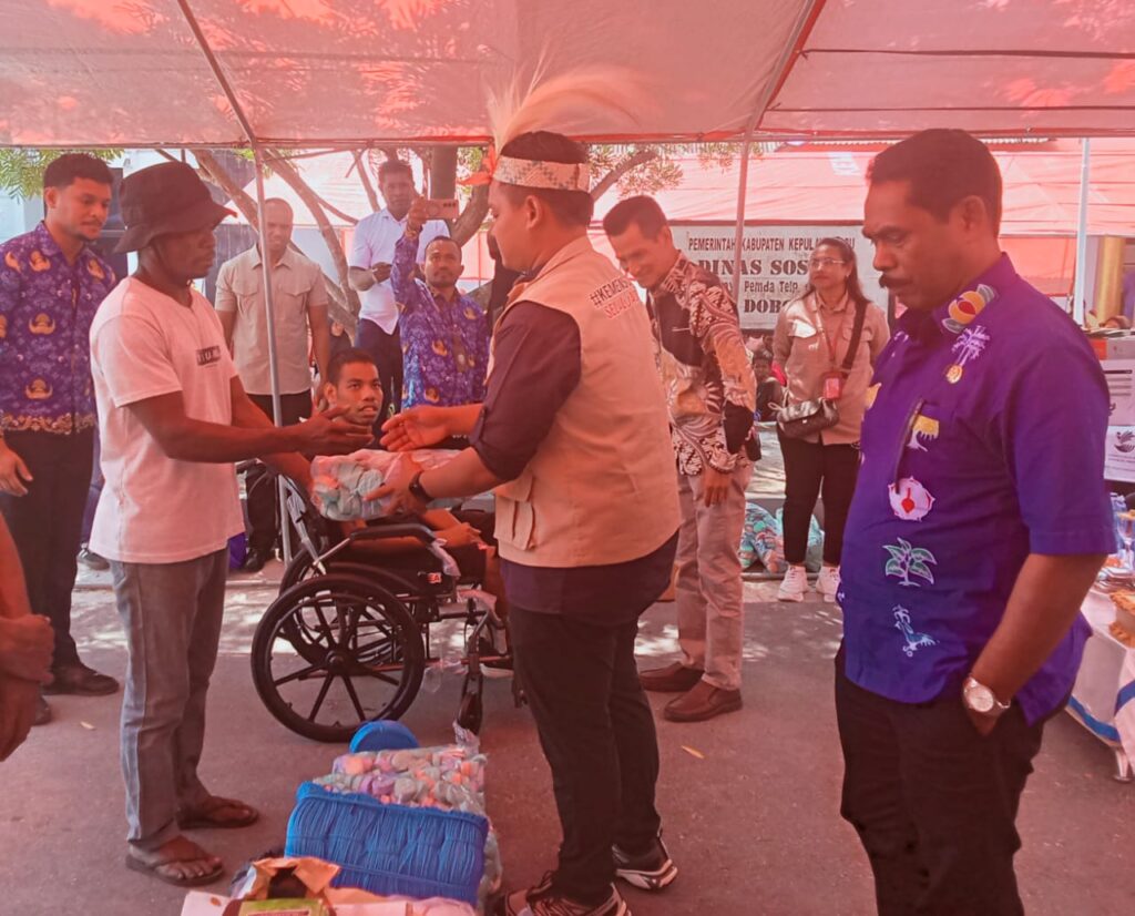 Mengenakan rompi krem dan hiasan kepala tradisional saat menyerahkan paket bantuan kepada warga di Dobo.
