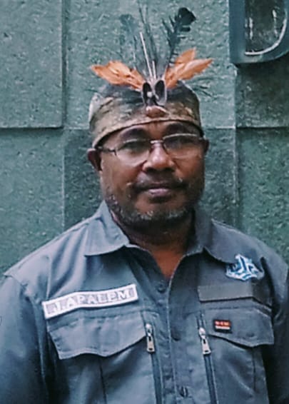 Potret seorang pria berseragam serikat atau organisasi dengan mahkota hiasan bulu khas Papua menatap ke depan.